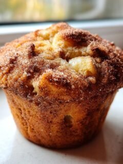 Close-up of a golden-brown Apple Cinnamon Muffin with visible chunks of apple and a cinnamon-sugar topping.