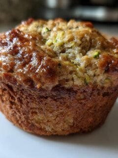 Close-up of a single, golden-brown Banana Zucchini Muffin on a white plate, showing its moist texture.