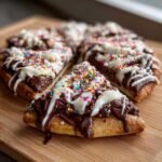Close-up of sweet chocolate pizza slices topped with chocolate spread, white icing, and colorful sprinkles.