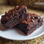 Two rich, dark chocolate brownies studded with chocolate chips on a white plate.