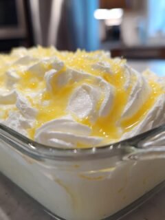 A close-up of a glass dish filled with creamy white Cream Cheese Gelatin dessert, topped with swirls of whipped cream and a drizzle of yellow sauce.