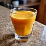 A close-up of a glass containing an Energizing Ginger Turmeric Shot, showing its vibrant orange color and frothy top.