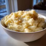 A close-up of a white bowl filled with creamy, flavorful roasted garlic mashed cauliflower, seasoned with pepper.