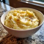 A close-up of a bowl filled with Irresistible Roasted Garlic Mashed Potatoes, topped with melted butter and spices.