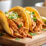Close-up of three irresistible slow cooker chicken tacos filled with shredded chicken and cilantro on a wooden board.