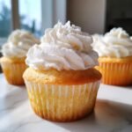 Close-up of an irresistible vanilla cupcake topped with swirls of fluffy white frosting, with more cupcakes in the background.