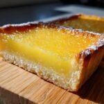 A close-up of a slice of moist lemon tart on a wooden board, dusted with powdered sugar.