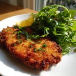 A golden-brown, crispy Pork Milanese served with a fresh arugula salad and a lemon wedge.