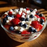 A close-up of a festive Red White and Blue Cheesecake Salad in a glass bowl, topped with fresh strawberries, blueberries, and mini marshmallows.