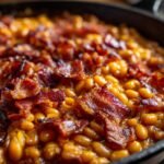 Close-up of Ultimate Baked Beans in a cast iron skillet, topped with crispy bacon.