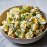 A close-up of Ultimate Delicious Potato Salad in a bowl, featuring creamy dressing, chunks of potato, sliced hard-boiled eggs, and fresh dill.