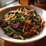 Close-up of a plate of glossy Beef Chow Fun noodles mixed with slices of beef and bright green scallions.