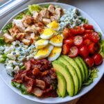 A vibrant, fully loaded Cobb Salad featuring rows of grilled chicken, blue cheese, hard-boiled eggs, bacon, avocado, and tomatoes.