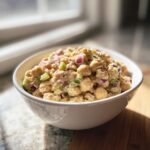 A white bowl filled with creamy Chickpea Salad mixed with diced red onion and green celery, set near a sunlit window.