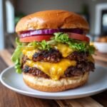 Close-up of a tall, juicy double patty California Burgers stacked high with melted cheese, lettuce, tomato, onion, and pickles.