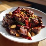 A close-up of glossy, saucy Easy Kung Pao Beef pieces mixed with roasted peanuts and dried red chilies on a white plate.