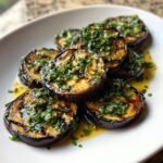 Close-up of perfectly Grilled Eggplant With Garlic Vinaigrette slices drizzled with oil and topped with fresh herbs.