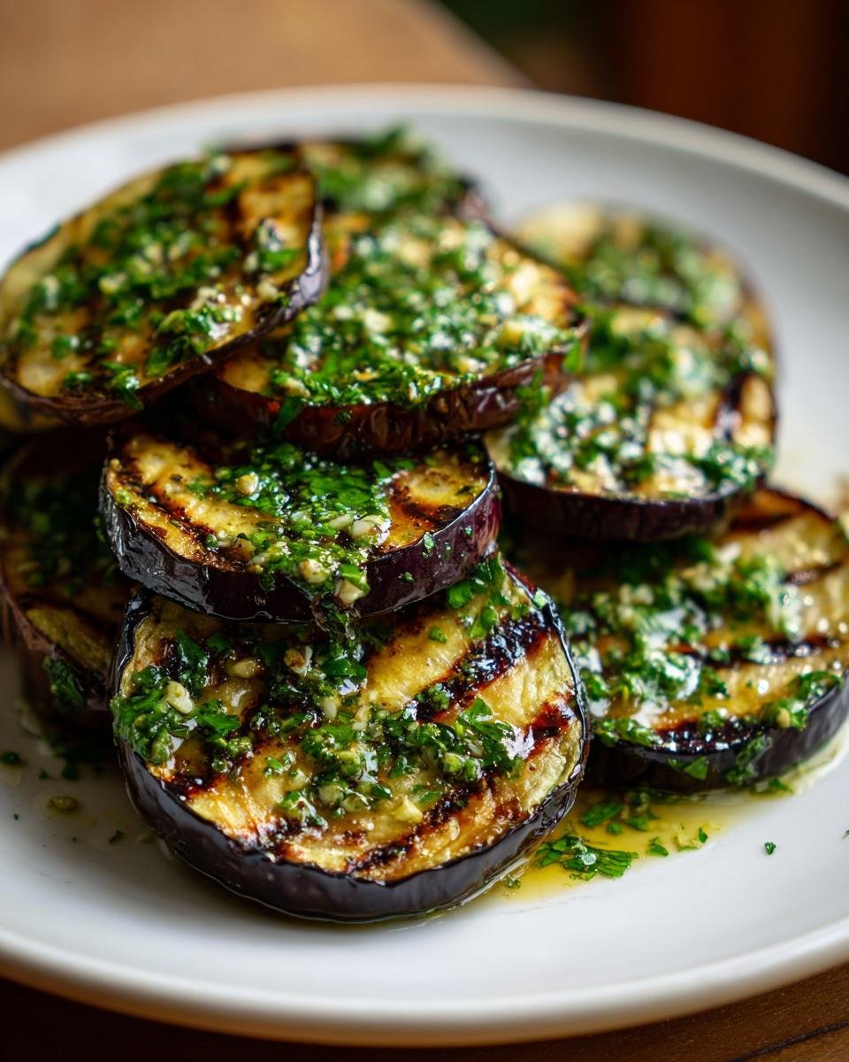 Close-up of stacked, charred slices of Grilled Eggplant With Garlic Vinaigrette drizzled with herbs.
