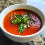 A close-up of a bowl of vibrant Homemade Tomato Soup With Fresh Tomatoes, garnished with fresh basil and black pepper.