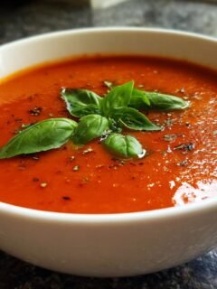 A close-up of a white bowl filled with vibrant Homemade Tomato Soup With Fresh Tomatoes, garnished with basil leaves and herbs.