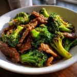 Close-up of Irresistible Beef And Broccoli Stir Fry in a white bowl, showing tender beef strips and vibrant broccoli florets coated in a glossy sauce.