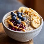 A close-up of an Irresistible Peanut Butter Banana Overnight Oats bowl topped with blueberries, jam, peanut butter, chia seeds, and banana slices.