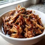 A close-up of shredded, tender Irresistible Slow Cooker BBQ Chicken coated in rich BBQ sauce, garnished with fresh herbs.