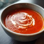 A close-up of a bowl of Irresistible Tomato Soup, topped with a swirl of cream and black pepper.