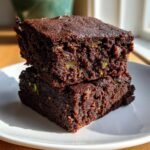 Two rich, dark chocolate Zucchini Chocolate Brownies stacked on a white plate, showing flecks of green zucchini.
