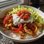 A close-up of a bowl filled with white rice, seasoned chicken, peppers, topped with lettuce, sour cream, and salsa for Ranch Chicken Fajita Bowls.