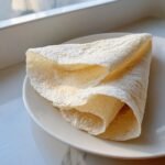 A lightly browned, folded Egg White Wraps resting on a white plate near a bright window.