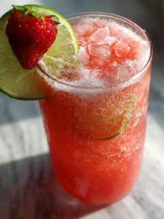 A tall glass filled with a bright red, icy Strawberry Beer Margaritas, garnished with a lime wheel and fresh strawberry.