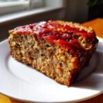A close-up of a thick slice of Air Fryer Meatloaf topped with a shiny red ketchup glaze, served on a white plate.
