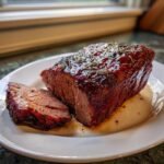 A thick slice of glazed Pork Roast, topped with rosemary, resting on a white plate with pan juices.