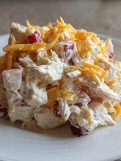A mound of creamy Apples Cheddar Chicken Salad topped with shredded cheddar cheese, served on a white plate.