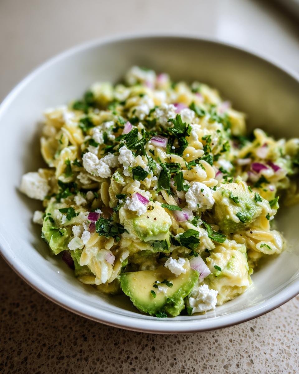 A close-up view of a bowl filled with vibrant Avocado Feta Orzo Salad, featuring creamy avocado chunks, feta cheese, and herbs.