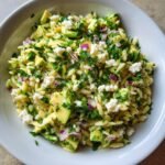 A close-up, overhead view of a vibrant Avocado Feta Orzo Salad featuring orzo pasta, chunks of avocado, feta cheese, and red onion.