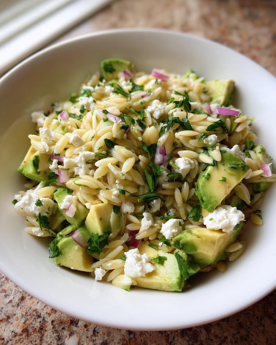 A close-up view of a white bowl filled with fresh Avocado Feta Orzo Salad, featuring pasta, avocado chunks, feta cheese, and red onion.