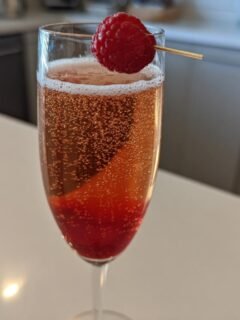 A tall flute glass filled with a sparkling pink Chambord Bellini, garnished with a fresh raspberry on a pick.