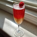 A layered Chambord Bellini cocktail in a flute glass, featuring red and gold layers, garnished with a fresh raspberry.