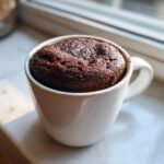 A rich, dark Chocolate Mug Cake risen perfectly inside a white ceramic mug, set near a window.