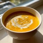 Close-up of a bowl of bright orange Creamy Carrot Soup topped with a swirl of cream and cracked black pepper.