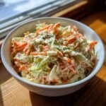 A close-up of creamy Coleslaw featuring shredded cabbage and carrots in a white bowl, highlighted by natural sunlight.