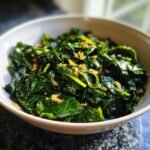 A close-up of vibrant, wilted Garlic Butter Collard Greens topped with browned garlic pieces in a white bowl.
