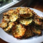 A close-up of crispy, golden Garlic Parmesan Baked Zucchini Chips piled high on a white plate.