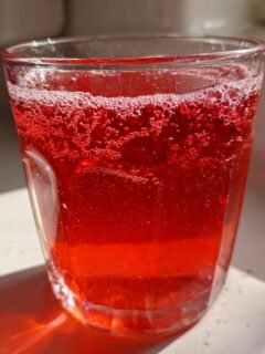 A glass filled with bright red, fizzy Homemade Strawberry Juice sitting in sunlight.