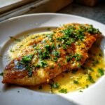A close-up of a golden-brown Chicken Paillard fillet served on a white plate, drenched in a bright lemon butter sauce and topped with fresh parsley.