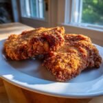 Two perfectly golden brown, crispy Fried Pork Chops resting on a white plate near a bright window.