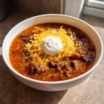 A close-up of a white bowl filled with rich Taco Soup, topped with shredded cheese, sour cream, ground beef, and kidney beans.