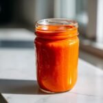 A glass mason jar filled with vibrant orange Homemade Catalina Salad Dressing, sitting on a white marble surface.